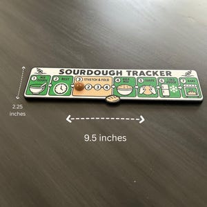 Puede incluir: Un tablero de seguimiento de masa madre rectangular que mide 5,7 cm por 24,1 cm. El tablero presenta siete pasos numerados para hacer pan: Mix Dough, Rest, Stretch & Fold, Bulk Rise, Shape, Cold Proof y Bake, con un peque&ntilde;o marcador en forma de pan.