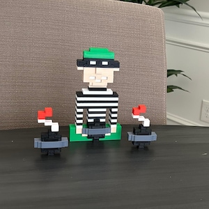 May include: A pixelated figurine of a burglar wearing a black and white striped shirt, black mask, and green hat. Two pixelated bombs with red fuses are on either side of the burglar. The figurine is on a green base.