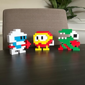 May include: Three pixel art figures standing in a row. The left figure is white and blue with a red accessory. The middle figure is yellow and red with small feet. The right figure is green, white, and red, featuring a blocky, jagged shape.