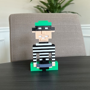 May include: A 3D printed figurine of a person wearing a black and white striped shirt, a green hat, and black sunglasses. The figurine is sitting on a green base.