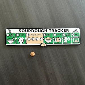 May include: A rectangular white and green sourdough tracker sign with black text and illustrations detailing seven steps: mix dough, rest, stretch and fold, bulk rise, shape, cold proof, and bake. A small, round, tan bread-shaped marker is included.