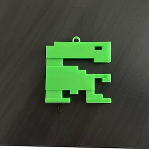 May include: A bright green, pixelated dinosaur ornament. The 3D-printed decoration features a loop at the top for hanging. The design is reminiscent of classic video game graphics, with a blocky, geometric aesthetic.