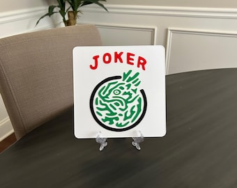May include: A white square sign with the word "JOKER" in red letters at the top. Below the word is a green and black circular design. The sign is displayed on a clear plastic stand, set on a dark gray table.