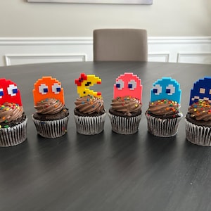 May include: Six chocolate cupcakes decorated with colorful sprinkles and topped with Pac-Man character figurines. The figurines are in various colors, including red, orange, yellow, pink, blue, and dark blue.