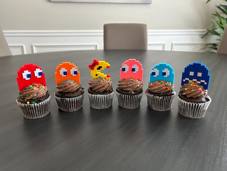 May include: Six chocolate cupcakes decorated with colorful sprinkles and topped with Pac-Man character figurines. The figurines are in various colors, including red, orange, yellow, pink, blue, and dark blue.
