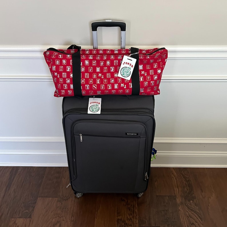 May include: A black rolling suitcase with a red patterned tote bag on top. The tote bag has a repeating pattern of white and red squares. Both bags have a tag that says "Joker".