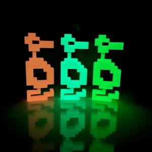 May include: Three pixelated figures, one orange, one green, and one teal, stand on a black surface. The figures are reflected in the surface.