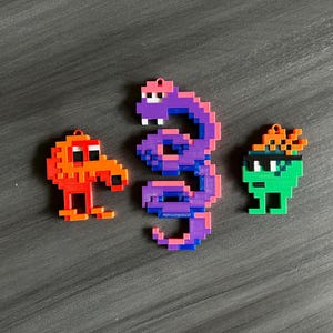 Puede incluir: Tres adornos de arte de píxeles, una serpiente morada, un personaje verde con una diadema naranja y un personaje naranja con un pico rojo.