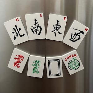 Puede incluir: Un conjunto de ocho imanes rectangulares con forma de fichas de mahjong de superficie blanca. La fila superior muestra caracteres direccionales con letras rojas N, S, E, W. La fila inferior presenta motivos de dragones rojos y verdes.