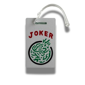 May include: A rectangular, light gray luggage tag with the word "JOKER" in red letters. Below the text is a black circle with a green, stylized design. A clear plastic loop is attached at the top.