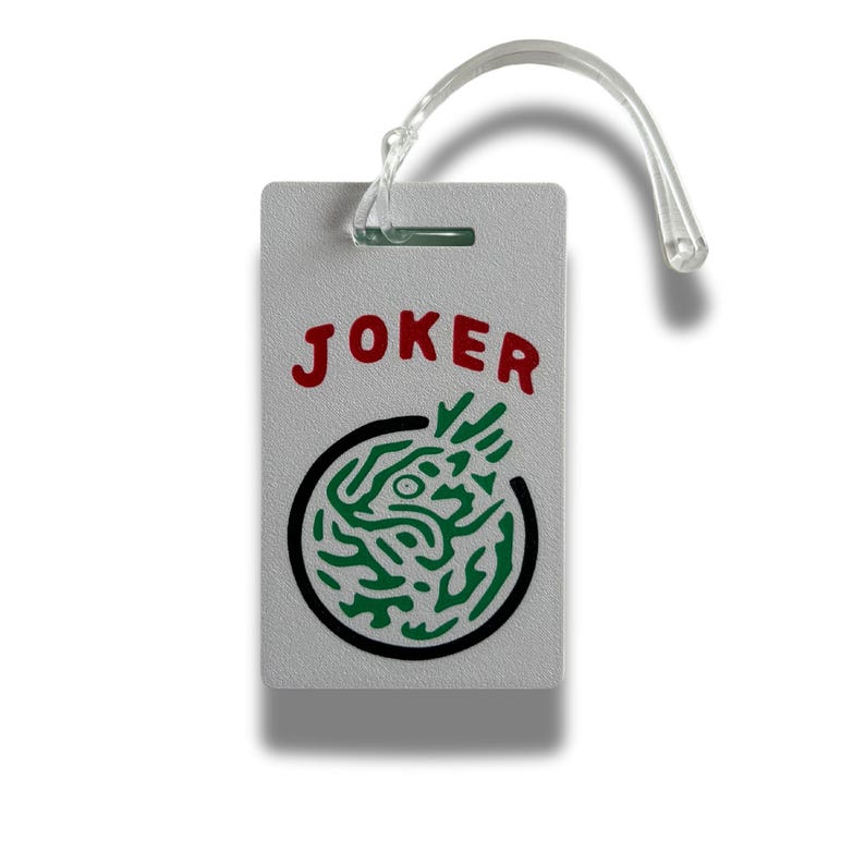 May include: A rectangular, light gray luggage tag with the word "JOKER" in red letters. Below the text is a black circle with a green, stylized design. A clear plastic loop is attached at the top.