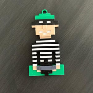 May include: A pixelated ornament depicting a cartoon burglar. The figure has a green hat, black mask, and a black and white striped shirt. The base and the loop are green. The figure is made of plastic.