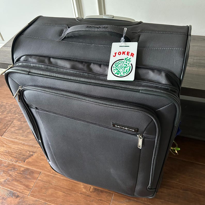 May include: A dark gray Samsonite rolling suitcase with multiple zippered compartments. A white tag with the word "JOKER" in red and a green design hangs from the handle. The suitcase is on a wooden floor.