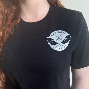 Może przedstawiać: Czarny T-shirt z białym nadrukiem przedstawiającym nietoperza z skrzyżowanymi mieczami i tekstem "BAT BOYS BOOTY CAMP".