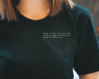 May include: Black crew neck t-shirt with white text on the upper left chest. The text reads: "Someone to lovers, dark haired and morally gray, dagger to the throat, only one bed, who did this to you?" A vintage silver camera is held in the lap.