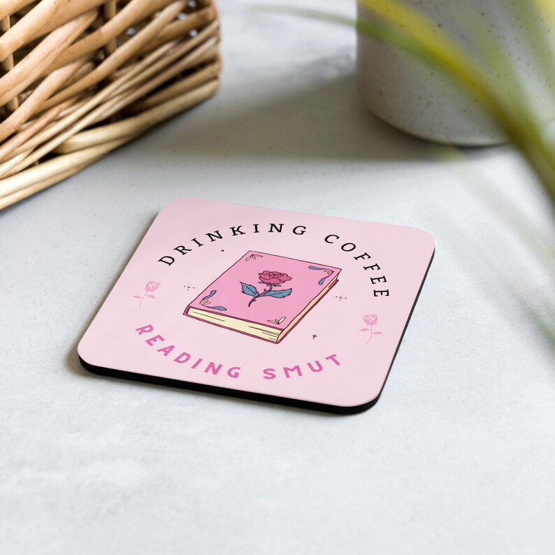 May include: Pink square coaster with black border featuring a book with a rose on the cover and the text "Drinking Coffee Reading Smut".