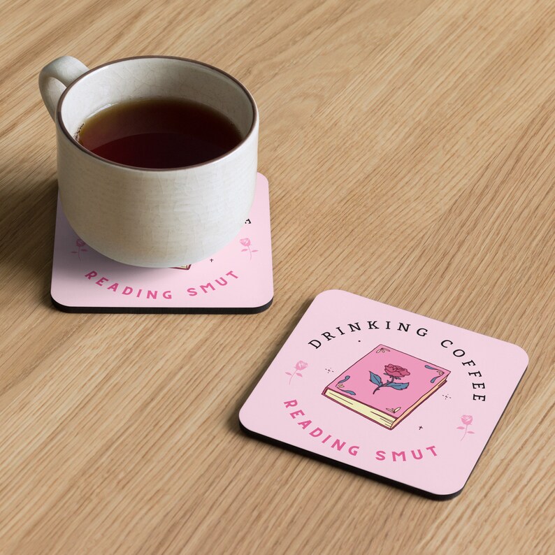 May include: Two pink square coasters with a book and rose design. The top coaster says "Drinking Coffee Reading Smut" and the bottom coaster says "Reading Smut".