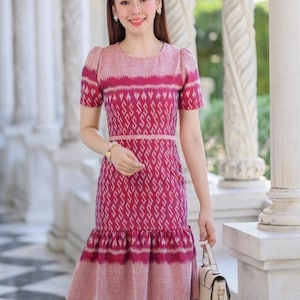 May include: A pink and white patterned dress with a ruffled hem and a matching belt. The dress is made of a lightweight fabric and has short sleeves. The model is holding a small beige handbag.