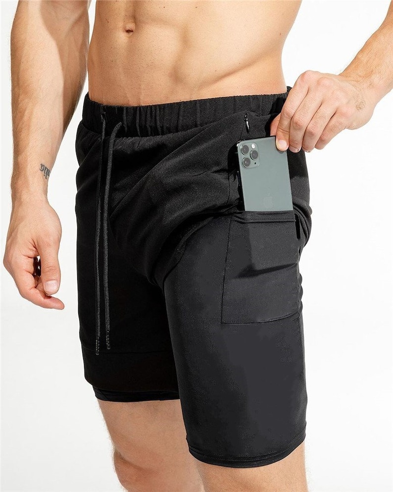May include: Black athletic shorts with a zippered pocket on the right side. The shorts have a drawstring waistband and a second layer of fabric underneath.