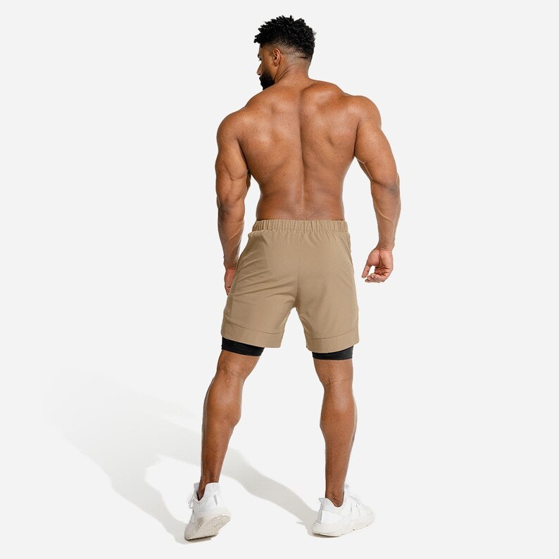 May include: A man wearing khaki athletic shorts with black inner shorts. He is standing with his back to the camera and wearing white sneakers.
