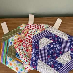 May include: Four quilted pot holders with different floral patterns. The pot holders are made of patchwork fabric in shades of green, red, blue, white, and yellow. Each pot holder has a loop for hanging.