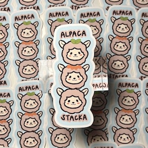 Op de afbeelding: Een verzameling Alpaca-stickers op een lichtblauwe achtergrond. De stickers hebben een schattig cartoon alpaca-ontwerp met een stapel van drie alpaca's. Het woord "ALPACA" staat bovenaan de stickers en "STACKA" onderaan.
