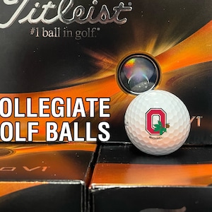 May include: A white golf ball with a collegiate logo, resting in front of a box of golf balls. The box is black and orange with the text "Titleist" and "#1 ball in golf." The text "COLLEGIATE GOLF BALLS" is also visible.