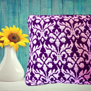 May include: A purple and white crocheted pillow with a damask pattern. The pillow is sitting on a white surface in front of a teal blue wooden wall.