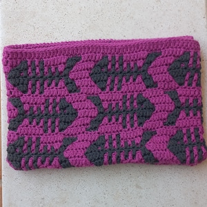 Op de afbeelding: Een gehaakte clutch met een roze achtergrond en een zwart geometrisch patroon. Het patroon bestaat uit herhalende vormen die lijken op visgraten.