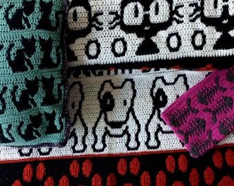May include: A crocheted blanket with a black and white cat design, a black and white dog design, and a red paw print design. The blanket is made with a variety of colors, including black, white, red, and green.