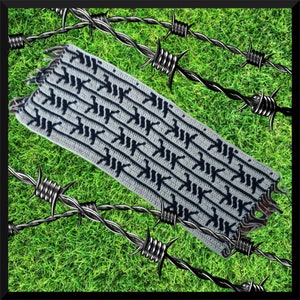 Könnte beinhalten: Ein weißer Häkel-Schal mit einem schwarzen, sich wiederholenden Muster des Buchstabens "K". Der Schal liegt auf Stacheldraht.