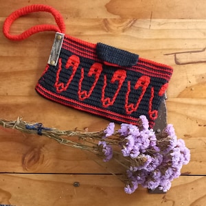 May include: A black and red crocheted purse with a red handle and a small pocket on the front. The purse is decorated with a pattern of safety pins.