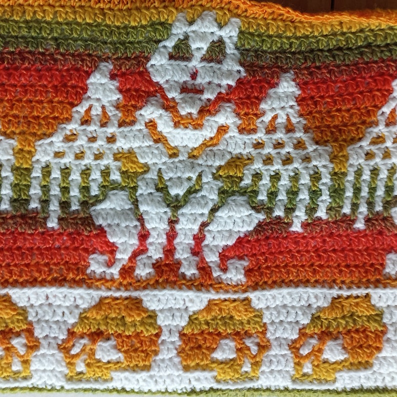May include: A crocheted blanket with a white, orange and green pattern. The pattern features a repeating design of skeletons holding hands.