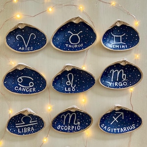 May include: Twelve hand-painted seashell dishes, each featuring a different zodiac sign in white script against a dark blue, starry background. The shells have a natural, beige rim and are arranged on a light-colored surface.
