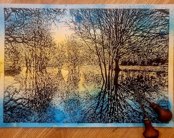 Puede incluir: Una impresión de linóleo en blanco y negro de una escena de bosque inundado con árboles reflejados en el agua. La impresión está sobre un fondo azul y amarillo con dos herramientas de talla de madera en primer plano.