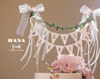 May include: White wedding cake topper with a banner that reads "JUST MARRIED" in black lettering. The banner is strung between two white ribbon bows with green leaves. The cake topper is made by HANA forest.