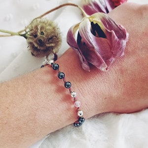 May include: A beaded bracelet featuring dark metallic hematite beads, polished silver-tone beads, and a single translucent light pink bead, all strung together on a thin, dark pink cord. The beads are arranged in a repeating pattern along the cord.