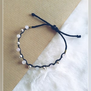 May include: A black cord bracelet featuring alternating pale pink beads and metallic silver-toned beads. The beads are secured along the cord with thin gold-toned wire wraps. The bracelet includes an adjustable sliding knot closure with two loose ends.