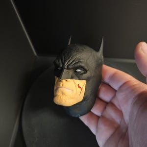 May include: A black and yellow Batman head sculpt with a red scratch on the cheek. The sculpt is made of plastic and has a detailed face.