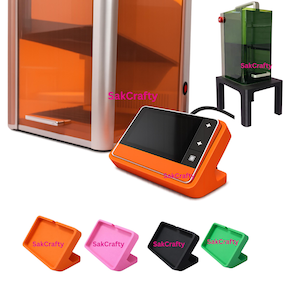 May include: An orange 3D printer control panel with a black screen and buttons, connected to a 3D printer. Additional colored stands in orange, pink, black, and green are displayed. The text "SakCrafty" is visible.