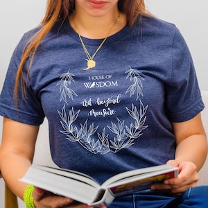 May include: A woman wearing a navy blue t-shirt with a white design that reads "House of Wisdom, wit beyond measure". She is holding a book open in her hands.