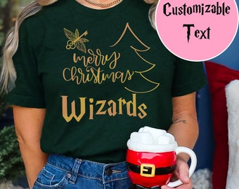 May include: Dark green t-shirt with gold text that reads "merry Christmas Wizards" and a stylized Christmas tree and key graphic. A pink circle with the words "Customizable Text" is in the upper right. A Santa mug is held in the hand.