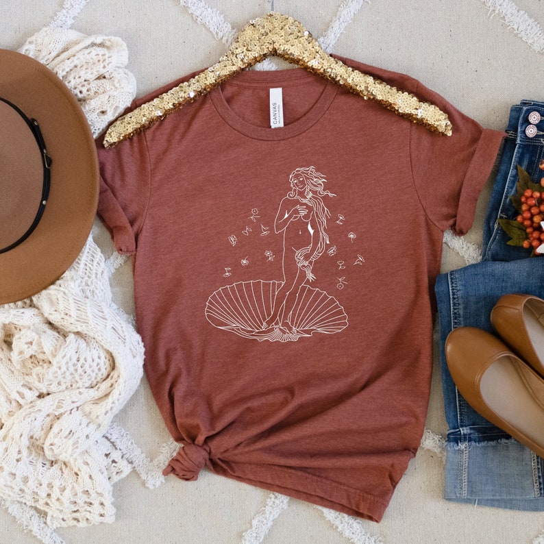 May include: A rust-colored t-shirt with a white outline of the Birth of Venus painting. The painting depicts a woman standing on a seashell with her hair flowing behind her.