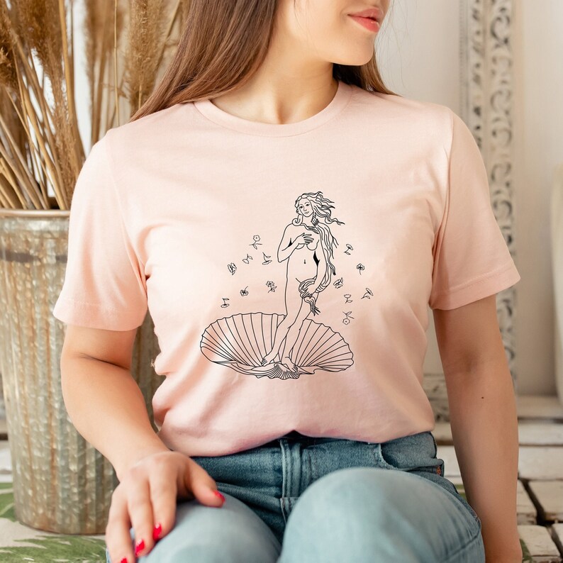 May include: A pink t-shirt with a black line drawing of the Birth of Venus on the front. The drawing includes a woman standing on a seashell with flowers around her.