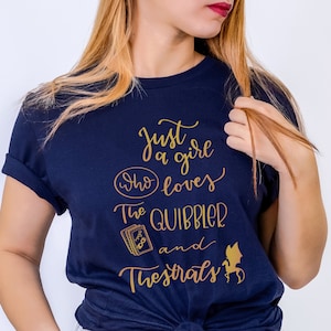 May include: A navy blue t-shirt with gold lettering that reads "Just a girl who loves The Quibbler and Thestrals". The shirt features a graphic of a book and a Thestral.