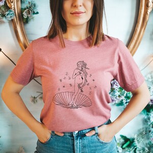 May include: A pink t-shirt with a black line drawing of the Birth of Venus, a famous Renaissance painting by Sandro Botticelli. The drawing depicts a nude woman standing on a seashell, with her hair flowing in the wind.