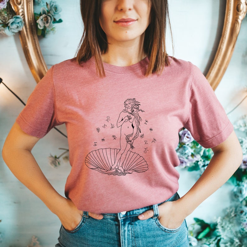 May include: A pink t-shirt with a black line drawing of the Birth of Venus, a famous Renaissance painting by Sandro Botticelli. The drawing depicts a nude woman standing on a seashell, with her hair flowing in the wind.