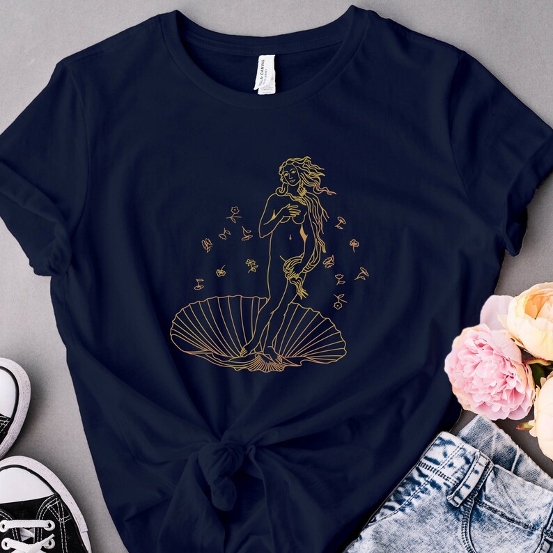 May include: A navy blue t-shirt with a gold outline illustration of the Birth of Venus, a famous Renaissance painting. The illustration depicts a woman standing on a seashell with flowers falling around her.