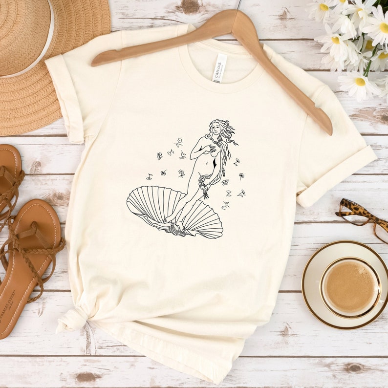 May include: A white t-shirt with a black line drawing of the Birth of Venus on the front. The drawing shows a woman standing on a seashell with her arms crossed over her chest. The woman has long flowing hair and is surrounded by small circles.