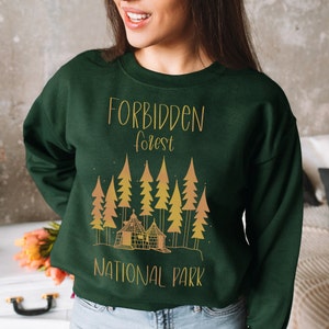 May include: A dark green sweatshirt with a gold graphic design of a cabin in a forest. The text "Forbidden Forest National Park" is printed on the sweatshirt.